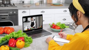 Woman in home kitchen study online video call webcam laptop listen chef teacher, writes in notebook. Man food blogger in computer screen tells teaches housewife remote culinary lesson, slices cucumber