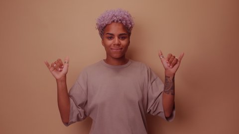Friendly looking positive African American woman blonde haircut shows shaka sign gestures indoor being in high spirit, wears fashionable t shirt isolated over beige background