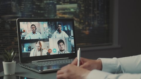 Computer monitor with video conference of multiracial doctors' leaders. Hands writing notes in front.