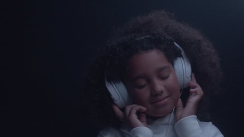 Portrait of happy smiling african american child girl listening music wearing headphones, moving in dynamic light indoors close up. Having fun, dancing, enjoying cool relax using modern sound device