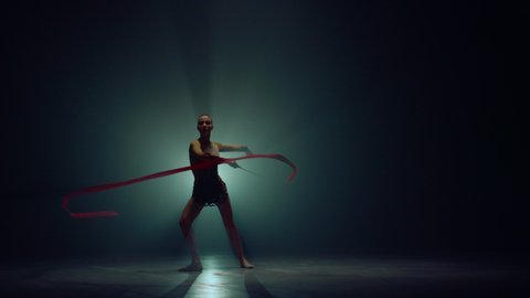 Young sportswoman spinning with gymnastic ribbon in dark room. Flexible woman practicing rhythmic gymnastics indoors. Beautiful female gymnast moving hand with red tape in spotlight background.