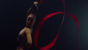 Beautiful girl exercising with gymnastic ribbon in dark space. Young woman training with flying tape in hand indoors. Pretty rhythmic gymnast posing for camera inside.