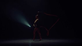 Slim woman performing with gymnastic ribbon in spotlight background. Young gymnast twirling hand with flying tape in dark space. Flexible sportswoman dancing indoors. Rhythmic gymnastics concept. 