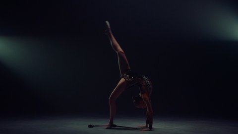 Flexible girl doing somersault with sports equipment in spotlight background. Slim sportswoman throwing maces in dark space. Sporty woman practicing rhythmic gymnastics indoors.