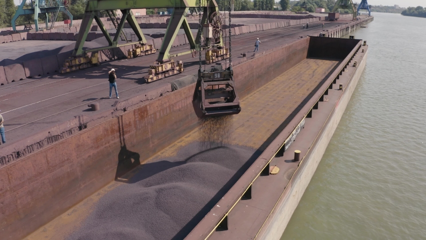 Drone aerial shot of loading ore at the port. A loader pours iron ore onto a barge. Several unrecognizable men in helmets are standing on the pontoon. One of them is filming the loading process. Royalty-Free Stock Footage #1076349710