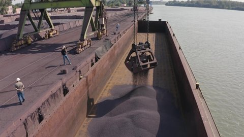 Drone aerial shot of loading ore at the port. A loader pours iron ore onto a barge. Several unrecognizable men in helmets are standing on the pontoon. One of them is filming the loading process.