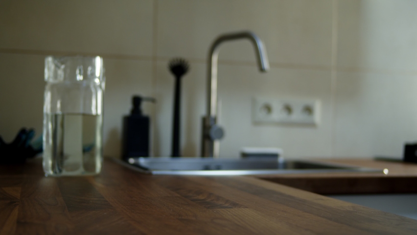 Closeup female hands pouring water into glass from jug at kitchen background. Unrecognizable person taking waterglass to drink at home. Unknown woman standing in modern apartment alone.