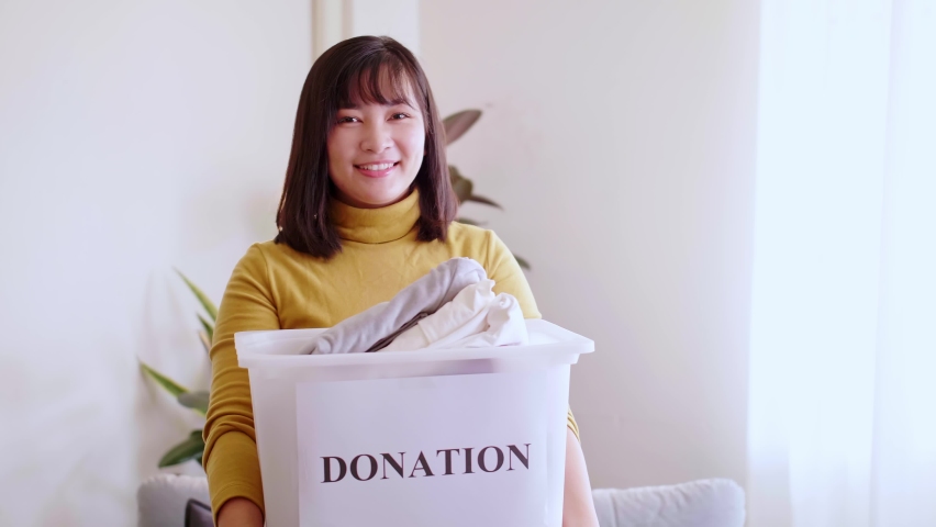 Portrait of young asian woman looking at camera holding box with clothes for donation. Royalty-Free Stock Footage #1076361953