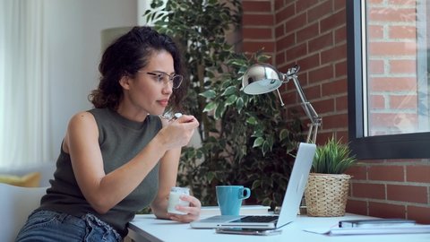 Video of beautiful young business woman working with laptop while eating yogurt in living room at home.