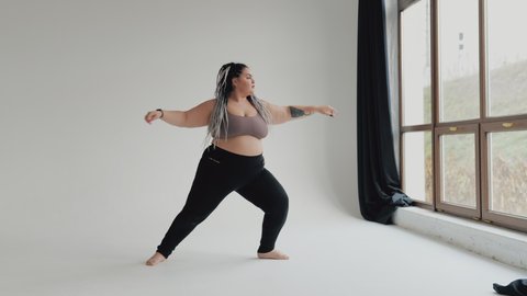 Happy body positive fat woman with dreadlocks doing yoga in the gym. Concept of natural diverse beauty and sport. Stout over weight female stretching isolated. Relaxation and tranquilty concept.