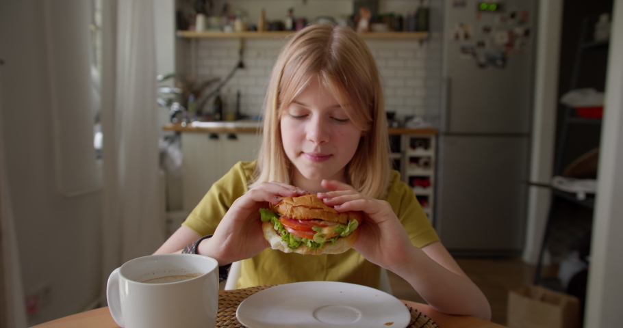 Lovely hungry teenager taking delicious fresh burger from plate, biting it with appetite and enjoyment, expressing positive emotions while having breakfast at home Royalty-Free Stock Footage #1076369540