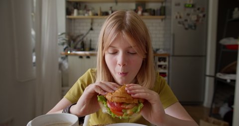 Lovely hungry teenager taking delicious fresh burger from plate, biting it with appetite and enjoyment, expressing positive emotions while having breakfast at home