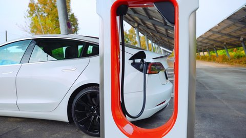 ROME, ITALY - APRIL 28, 2021: White premium Tesla car charging battery on supercharger station, autonomous electric car refilling energy from electric plug on parking lot. High quality 4k footage