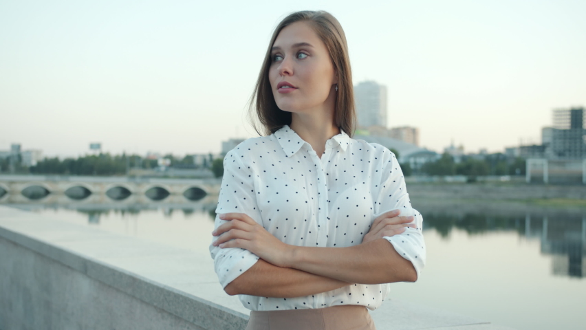 Portrait of ambitious young lady standing in embankment in modern city and turning to camera against beautiful urban background. People and business concept.