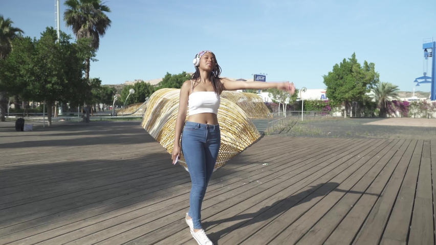 Dark-Skinned pretty girl with dreadlocks dancing in park during listen joyful music on headphones. Good vibes during good weather outdoors. Track shot.