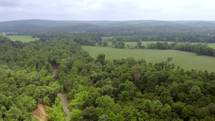 Pull back over trees and beautiful open pasture landscape with a stream in southern Missouri on a cloudy summer day.