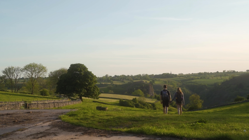 Couple walking hand-in-hand away from camera and then stopping to look at view of blue skies, green rolling hills and trees.