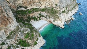 Small paradise cave beach between mountain cliffs in Spain with clear blue water and waves hitting the rocks.
Dron video panning.