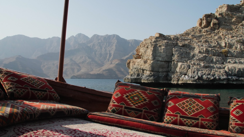 Tracking shot of ocean and fjords from the POV of a traditional Dhow boat moving through the water with colorful geometric pillows on a clear sunny day in Khasab, Musandam Oman. Royalty-Free Stock Footage #1076395292