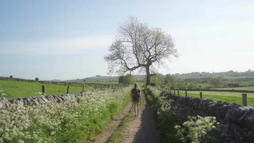 Slow-mo of blonde woman walking long a countryside path Ashbourne, Peak District, England. Green fields, blue skies, rolling hills, iconic drystone walls and trees in background.