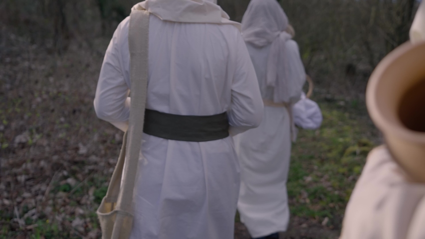 Overshoulder shot of three ladies walking on a path in a forest. They are wearing white white robes and they carry Jars with different ingredients. Royalty-Free Stock Footage #1076397740