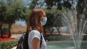 Slow motion video of a young European girl with a backpack wearing a mask standing and looking at the fountain.