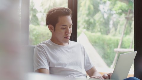 Young modern Asian man sitting by the window enjoying a relaxing moment working on a laptop in the coffee shop on a bright sunny day
