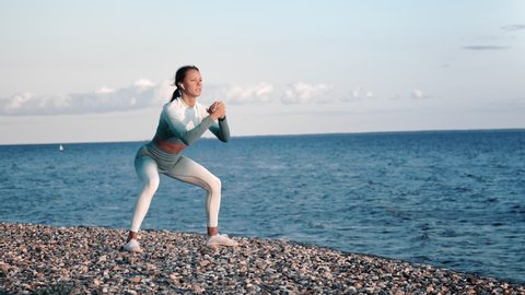 Athletic woman training on beach performing squat enjoying healthy lifestyle physical activity. Sports blonde female in sportswear doing outdoor training at sea landscape sunset shore slow motion