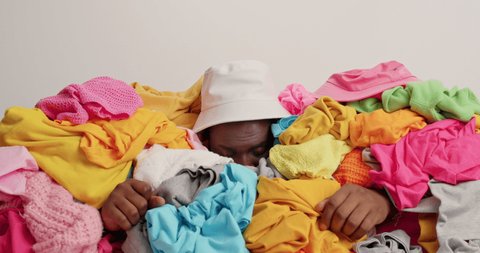 Overwhelmed dark skinned man buried in huge pile of multicolored laundry sticks head through unfolded clothes raises palms exclaims with indignant expression wears panama puzzled by real chaos
