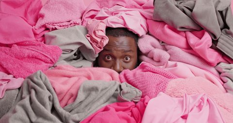 Dark skinned man covered with heap of clothes cluttered and tries to clean out wardrobe busy doing housework has surprised expression raises eyebrows. Messy closet. Human head stack in laundry