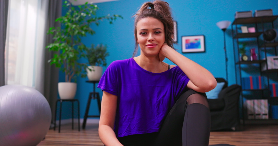 Pretty young woman dressed in fitness clothes chats on video chat with a personal trainer on a laptop while sitting on the wooden floor of an apartment. She looks into the web camera on laptop. Royalty-Free Stock Footage #1076433752