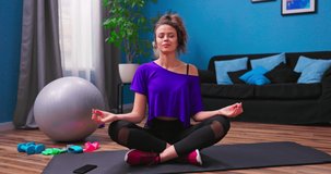 A pretty young woman dressed in fitness clothes meditates while sitting on a wooden floor in an apartment.