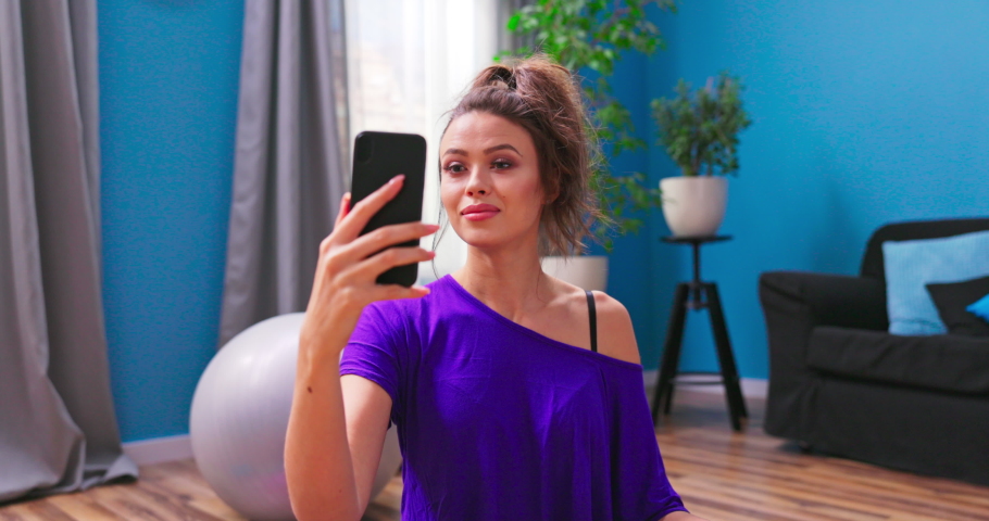 Pretty young woman dressed in fitness clothes chats on video chat with a personal trainer on cell phone while sitting on the wooden floor of an apartment. Royalty-Free Stock Footage #1076433773