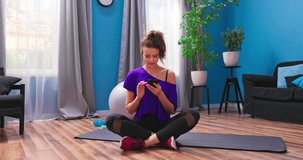 Beautiful young girl dressed in fitness clothes sits on the wooden floor of an apartment. Woman is using a smartphone, browsing content on the internet, searching video showing workout at home