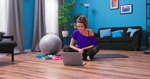 A pretty young woman dressed in fitness clothes chats on a video chat on a laptop while sitting on the wooden floor of an apartment.