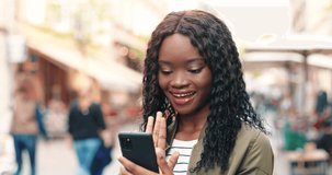 Waist up portrait view of a multiracial woman with long dark hair out and about in the city streets during the day, looking at the smartphone and waving with her hand to her friends. Technology and