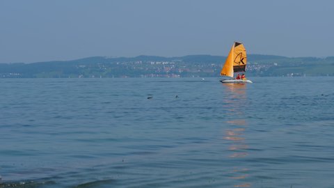 sailboat with orange sail near harbor Altnau. German shore at horizon. lake Bodensee, CH Switzerland. 20th July 2021