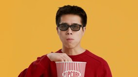 Asian Guy Wearing 3D Glasses Watching Movie And Eating Popcorn With Blank Face Expression Posing Over Yellow Studio Background, Looking At Camera. Fun And Entertainment, Cinema Concept