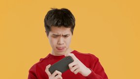 Chinese Young Man Using Phone Playing Game Online Standing Over Yellow Studio Background. Mobile Gaming, Fun And Entertainment Application Concept. Slow Motion