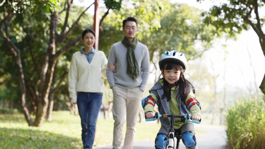 Little asian girl riding bike with full protective gears outdoors in park while parents watching from behind | Shutterstock HD Video #1076468834