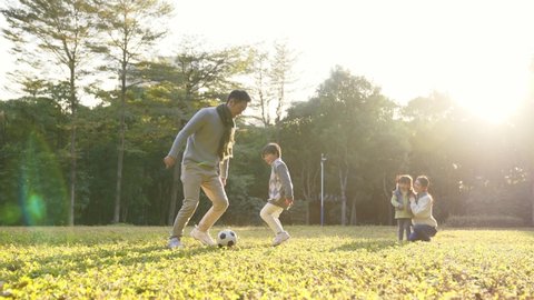 asian father playing soccer with son outdoors in park