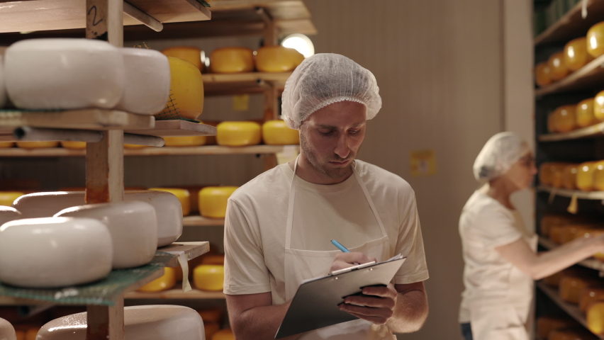 Male technologist in uniform walking at storage house with clipboard and examining cheese heads. Blur background of mature woman arranging cheese on wooden shelves. Craft production. Royalty-Free Stock Footage #1076469380