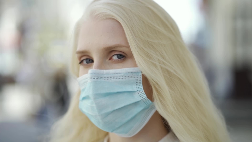 A blue-eyed blonde woman in a medical mask stands on the central street of a big city and looks into the camera. Coronavirus epidemic, personal protective equipment, quarantine. Royalty-Free Stock Footage #1076471717