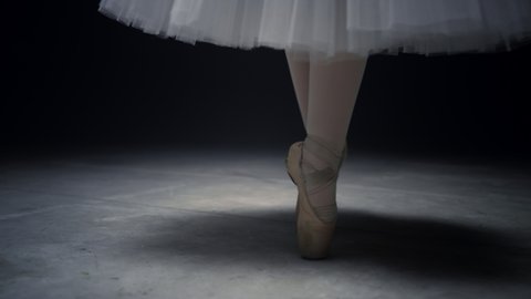 Closeup ballerina feet dancing in pointe shoes on stage. Ballet dancer legs standing on tiptoe in dark background. Unknown woman performing classic dance in pointe shoes indoors.