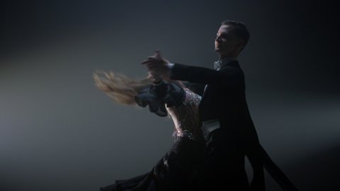 Full length of young dance couple twirling around on stage. Beautiful ballroom partners dancing indoors. Professional dancers performing valse in spotlights background.