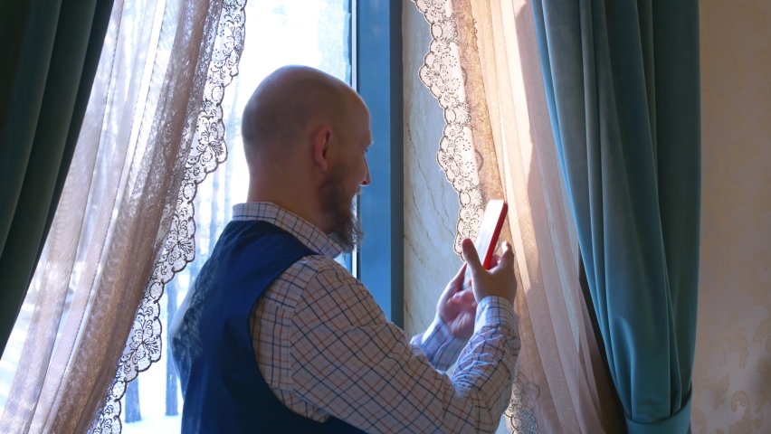 A man with a bald head and a stylish beard is standing next to the window, in a plaid shirt and a blue classic waistcoat, taking a selfie. The window is hung with blue curtains with patterned tulle.