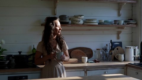 A pretty Asian young woman in a dress is standing in the morning in the sunny kitchen, drinking hot coffee or tea and enjoying her time