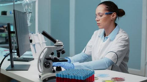 Researching specialist working in laboratory with chemical equipment. Woman using computer for dna examination while having microscope, petri dish, vacutainers and micropipette on desk