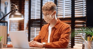 Young guy freelancer sitting alone in protective mask at modern apartment and working on laptop typing test emails for customers.