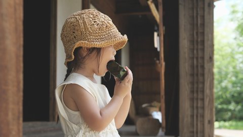 Asian little girl in straw hat sitting on the porch of old folk house and eating rice balls wrapped with seaweed. Swaying wind chime background. Summer season. Safe and healthy food for child growth